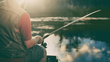 Person in einer Weste mit Netzeinsätzen sitzt beim Angeln im Boot auf einem ruhigen See im Sonnenlicht